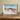 Framed artwork with a nature scene on a wall next to a wooden beam.