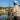 A man in a beige hunting uniform and wide-brim hat sits by a lake holding a shotgun loaded with ducks, with a rifle on a stand and a bucket nearby, against a natural outdoor background.