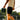 A man outdoors carrying a green and black portable speaker in one hand while holding a fishing rod in the other, wearing a cream t-shirt and black shorts.