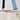 Close-up of a canvas tote bag with a light blue textured shoulder strap, a black shoulder strap, and gold-tone metal hardware against a white paneled background.