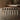 Six small cylindrical bottles with black caps arranged in a row on a dark wooden tabletop, photographed in a softly blurred indoor setting.