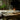 A rustic wooden dining table set for two with white plates, a clear glass carafe, two glasses, a white tablecloth, and a lit candle on a brass holder, in a dimly lit room with a stone fireplace.